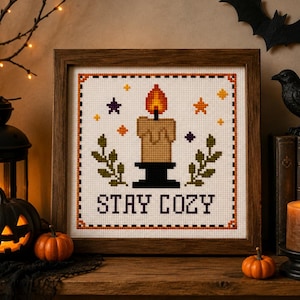 May include: A square cross-stitch embroidery in a dark wood frame featuring a tan candle with a bright orange flame, green leafy sprigs, and small colourful stars. The text STAY COZY is stitched in black letters at the bottom of the white fabric.