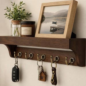 May include: A dark brown wooden wall shelf featuring five antique brass hooks. Three sets of keys hang from the hooks. A clear glass jar with a green plant, a brown candle jar labeled Cedar & Sandalwood, and a light wood picture frame sit on top.