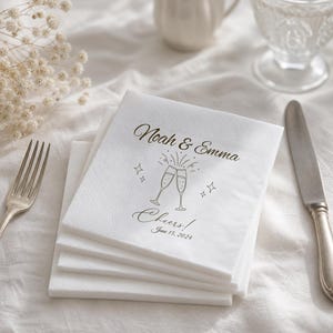Pode incluir: Uma pilha de guardanapos de papel branco com um design impresso em dourado de duas taças de champanhe a brindar, o texto "Noah & Emma", a palavra "Cheers!" e a data "June 15, 2024". Os guardanapos têm uma borda decorativa em relevo.