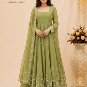 May include: An olive green floor-length dress with long sleeves, a square neckline, and a matching sheer dupatta. The hem features intricate silver embroidery. The text SuratTextileStudio SAREES | BLOUSES | KURTIS | LEHENGAS DESIGNED IN SURAT is visible.