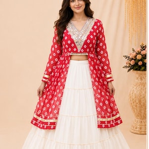 May include: A red and white patterned long-sleeved jacket with a V-neckline featuring mirror work, worn over a solid white tiered floor-length skirt. The jacket has a front opening and gold trim at the cuffs and hem. Text reads SuratTextileStudio.