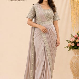 May include: A light taupe saree with a matching blouse featuring intricate silver embroidery on the sleeves, neckline, and waist belt. The garment includes a draped skirt section with a decorative border. Text reads SuratTextileStudio.