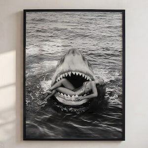 May include: A black and white framed wall art print depicting a large shark with its mouth wide open, revealing a person reclining inside the shark's jaws. The scene is set in rippling water, capturing a surreal composition with high contrast.