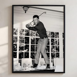 May include: A black and white framed wall art print depicting a person in a dark long-sleeved shirt and light trousers standing on a dining table. The person is holding a golf club in a swing position, with a large windowed wall in the background.