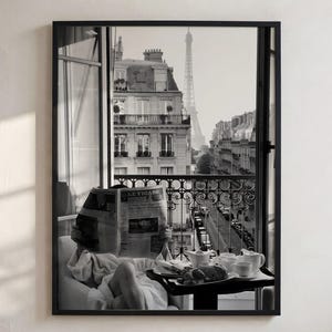 Könnte beinhalten: Ein schwarz-weiß gerahmter Wandkunstdruck, der einen Blick von einem Balkon auf eine Pariser Straße mit dem Eiffelturm in der Ferne zeigt. Eine Person im weißen Bademantel liest eine Zeitung an einem kleinen Tisch mit Frühstücksutensilien.