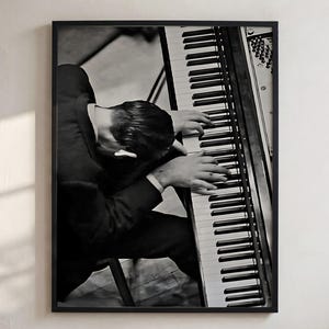 Könnte beinhalten: Ein schwarz-weiß gerahmter Wandkunstdruck, der eine Ansicht von oben auf eine Person in einem dunklen Anzug zeigt, die an einem Klavier sitzt. Die Person beugt sich mit gesenktem Kopf nach vorne, die Hände ruhen auf den schwarzen und weißen Klaviertasten. Die vertikale Komposition zeigt den Musiker und das Instrument von oben in einem monochromatischen Ton.