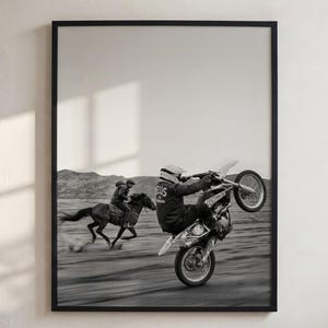 May include: A black and white framed wall art print depicting a person riding a dirt bike on a wheelie alongside a person riding a horse across a desert landscape. The rider wears a helmet and a jacket with the text DEUS EX MACHINA on the back.