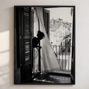 May include: A black and white framed wall art print depicting a person standing at an open window with wooden shutters. A sheer white curtain blows inward. The view through the window shows a narrow street with buildings and a metal balcony railing.