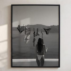 May include: A black and white framed wall art print depicting a person standing before a body of water, facing a clothesline where several swimsuits and bikini pieces are hanging from clips against a distant mountainous landscape background.