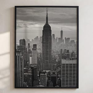 May include: A black and white framed wall art print featuring a high-angle cityscape of New York City. The central focus is the Empire State Building, surrounded by various skyscrapers and urban architecture under a cloudy sky.