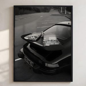 Op de afbeelding: Een zwart-wit ingelijste print met de achterkant van een donkere sportwagen op een parkeerplaats. Twee open pizzadozen en drankblikjes liggen op de achterspoiler van de auto. De kentekenplaat van het voertuig toont de tekst GG-627-NX.