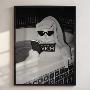 May include: A black and white framed wall art print featuring a cat wearing dark sunglasses and a towel on its head while sitting in a bathtub. The cat is holding an open book with the text HOW TO BE RICH clearly visible on the front cover.