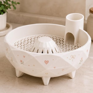 May include: A cream-colored, faceted, bowl-shaped dish featuring a raised central dome, a cylindrical side attachment, and a removable lattice insert. The exterior is decorated with small pink hearts, bows, and floral motifs on its geometric surface.