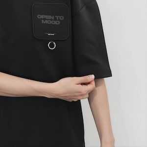 May include: A black short-sleeved t-shirt featuring a square chest pocket with rounded corners. The pocket displays the text "OPEN TO MOOD" in dark lettering and includes a silver-toned metal ring attached to the bottom edge of the pocket.