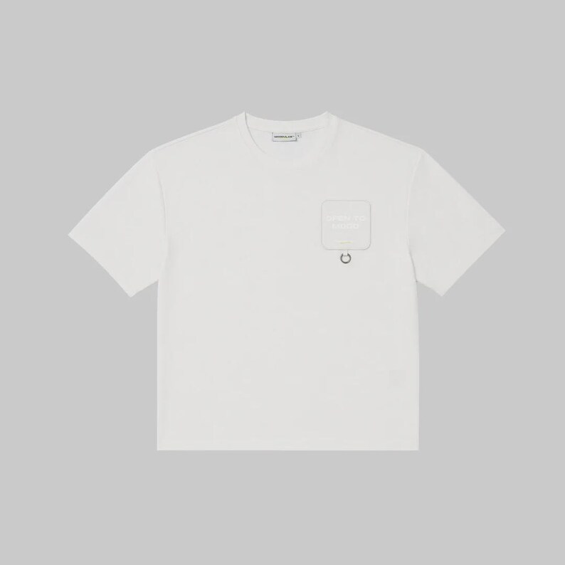 May include: A short-sleeved white t-shirt featuring a crew neckline and a square patch pocket on the left chest. The pocket includes a small metal ring attached at the bottom edge and subtle text printed on the surface of the pocket fabric.