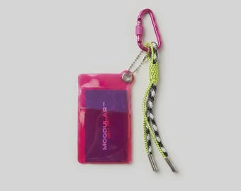 May include: A translucent pink plastic card holder featuring a purple insert with the text MODULAR WEAR SYSTEM. It is attached to a pink metal carabiner and a neon yellow and black patterned cord with silver-toned metal aglets at the ends.