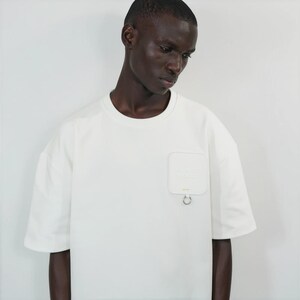 May include: An off-white short-sleeved t-shirt featuring a crew neckline and a square chest pocket on the left side. The pocket includes a small silver metal ring attached to the bottom edge, providing a distinct structural detail to the garment.