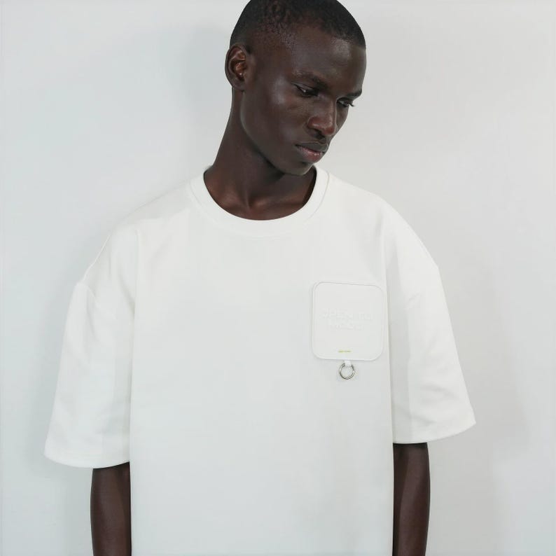 May include: An off-white short-sleeved t-shirt featuring a crew neckline and a square chest pocket on the left side. The pocket includes a small silver metal ring attached to the bottom edge, providing a distinct structural detail to the garment.
