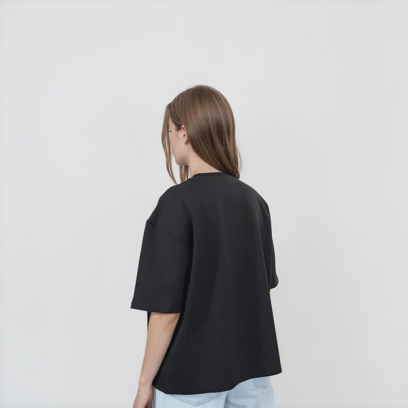 May include: A solid black short-sleeved t-shirt with a crew neckline and a loose, boxy fit. The garment is shown from the back, featuring plain fabric with no visible patterns, logos, or graphics, and is paired with light blue denim jeans.