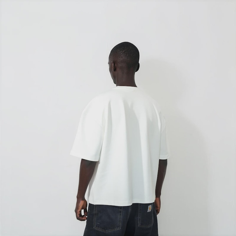 May include: A plain white short-sleeved t-shirt with a crew neckline and a loose, boxy fit. The garment is shown from the back, featuring a clean, solid-colored fabric design with no visible patterns, logos, or graphics on the rear surface.