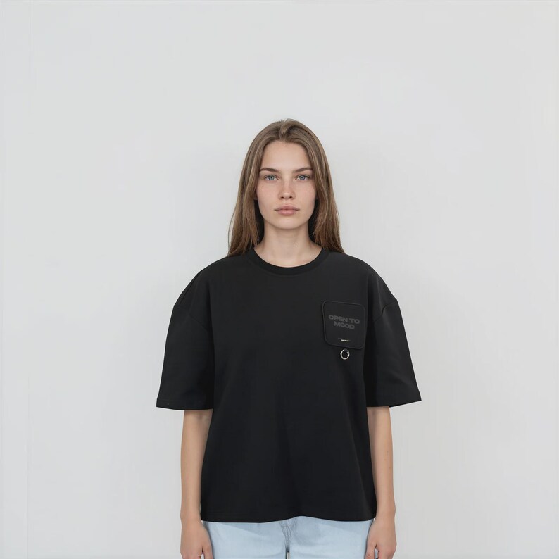 May include: A black short-sleeved t-shirt featuring a crew neckline and a square chest pocket on the left side. The pocket displays the text "OPEN TO MOOD" and includes a small silver-toned metal ring attached to the bottom edge of the pocket.