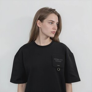 May include: A black short-sleeved t-shirt featuring a square patch on the left chest with the text "OPEN TO MOOD" printed in dark lettering. The patch includes a small metal ring attached to the bottom edge, adding a functional detail.