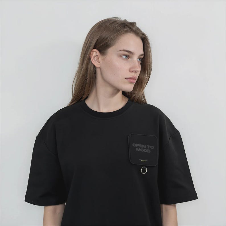 May include: A black short-sleeved t-shirt featuring a square patch on the left chest with the text "OPEN TO MOOD" printed in dark lettering. The patch includes a small metal ring attached to the bottom edge, adding a functional detail.