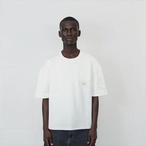 May include: A short-sleeved, off-white crew neck t-shirt featuring a single chest pocket on the left side. The pocket is adorned with a small, circular silver-toned metal ring detail attached at the bottom edge of the pocket fabric.