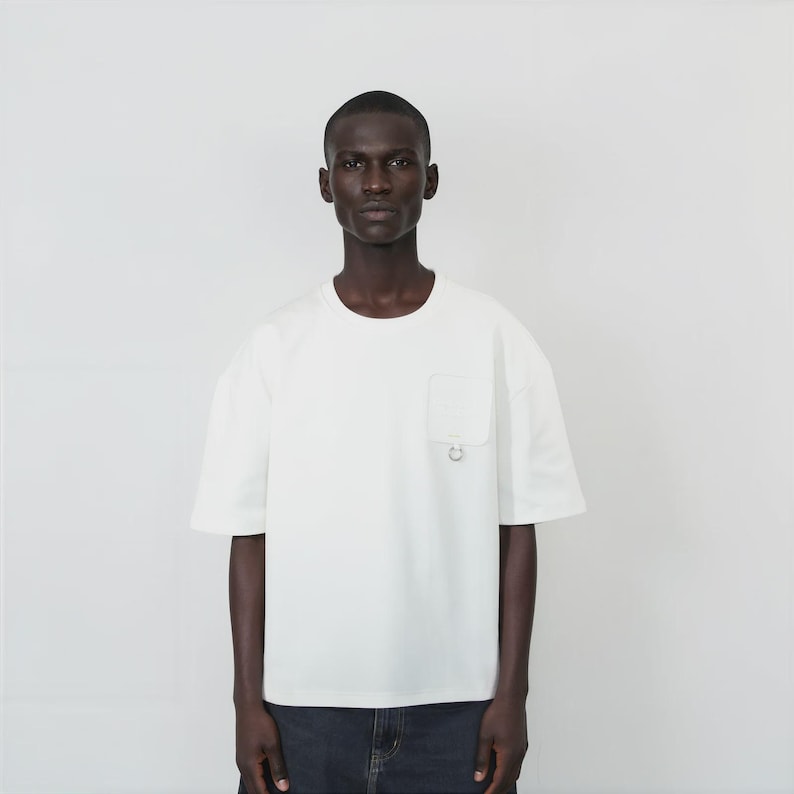 May include: A short-sleeved, off-white crew neck t-shirt featuring a single chest pocket on the left side. The pocket is adorned with a small, circular silver-toned metal ring detail attached at the bottom edge of the pocket fabric.