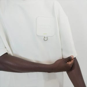 May include: An off-white short-sleeve t-shirt featuring a square chest pocket with rounded corners. The pocket displays the embossed text "OPEN TO MOOD" and a small silver metal ring attached to the bottom edge with a tiny lime green accent.