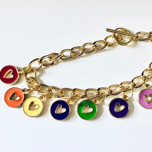 Puede incluir: Un brazalete de cadena dorada con un cierre de palanca y siete dijes coloridos. Cada dije es un disco redondo con un corazón recortado y está pintado en un color diferente: rojo, naranja, amarillo, azul, verde, morado y rosa.