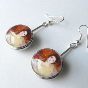 May include: A pair of silver earrings with round glass cabochons featuring a painting of a woman in a yellow dress. The earrings have a simple, elegant design.