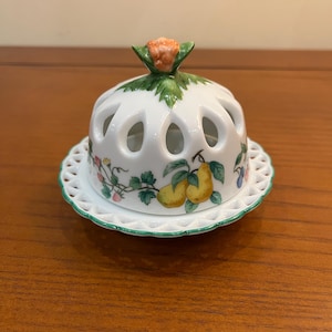 May include: A white porcelain butter dish with a domed lid featuring cut-out teardrop shapes and a fruit motif including yellow pears and green leaves. The lid is topped with a sculpted orange flower finial, and the base has a lattice edge.