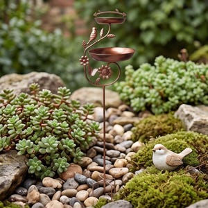 May include: A copper-coloured metal bird feeder stake featuring two circular bowls, a decorative branch with a small bird figure, and a sunflower accent. The structure is supported by a thin vertical rod with a four-pronged base for ground insertion.