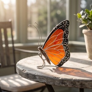 Può includere: Un ornamento a forma di farfalla monarca in vetro colorato con ali arancioni vivaci, venature nere e bordi a pois bianchi. La farfalla è sostenuta da un sottile supporto in filo metallico color argento che si curva per tenerla in verticale.