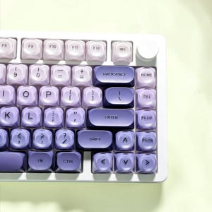 May include: A mechanical keyboard featuring a white frame and a gradient color scheme of purple and lavender keycaps. The keys display standard alphanumeric characters, function keys, and navigation controls, with a circular knob in the top right corner.