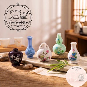 Könnte beinhalten: Eine Sammlung von fünf Miniatur-Keramikvasen in verschiedenen Formen und Farben, darunter eine dunkelviolette Vase mit weißen Blumen, eine blaue Vase, eine weiße Vase mit rosa Blumen, eine hellgrüne kürbisförmige Vase und eine weiße Vase mit blauen Blumen.