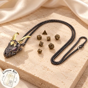 Puede incluir: Un collar de cadena de metal negro con un colgante de cabeza de dragón con cuernos dorados y ojos morados. La cabeza del dragón sostiene un pequeño dado poliédrico dorado. Seis dados poliédricos de color bronce están dispuestos cerca.
