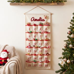May include: A cream-coloured fabric wall-hanging advent calendar with twenty-four pockets, each labelled with red numbers from 1 to 24. The name Amelia is embroidered in red at the top, and the pockets contain various small Christmas-themed items.