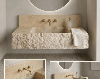 May include: A wall-mounted bathroom sink carved from beige natural stone with a rough, chiseled front edge. It features a smooth basin, a matching stone backsplash, and a brass faucet with two handles. A white towel hangs over the side.