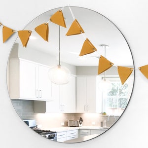 Może przedstawiać: Żółty, trójkątny baner w kolorze musztardowym z białym sznurkiem wiszącym na okrągłym lustrze. Baner odbija się w lustrze, które pokazuje kuchnię z białymi szafkami i lampą.