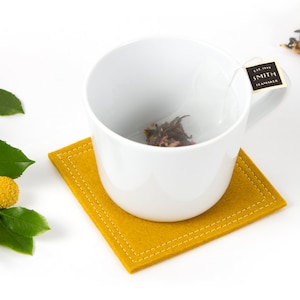 May include: A white ceramic mug with a tea bag inside, sitting on a yellow felt coaster. The tea bag has a label that reads "EST. 1900 SMITH TEA MAKERS".