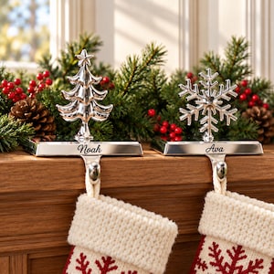 Könnte beinhalten: Zwei silberfarbene Metall-Strumpfhalter mit einem Weihnachtsbaum- und einem Schneeflocken-Motiv. Die Sockel sind mit den Namen Noah und Ava graviert. Die Halter stehen auf einem Holzsims und halten jeweils einen gestrickten Strumpf.