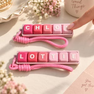 Könnte beinhalten: Zwei rechteckige Schlüsselanhänger mit einer Reihe von rosa Tasten im Stil mechanischer Tastaturen. Der obere Anhänger zeigt den Namen CHLOE in weißen Buchstaben, der untere den Namen LOTTIE. Beide haben eine geflochtene rosa Handschlaufe.