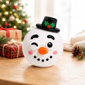 Könnte beinhalten: Eine runde, weiße Schneemann-Figur mit einem schwarzen Zylinder, der mit grünen Stechpalmenblättern und einer roten Beere verziert ist. Der Schneemann hat ein zwinkerndes Auge, ein offenes Auge, eine orangefarbene Karottennase und rosa Wangen.