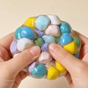 May include: A clear, flexible, circular sensory toy filled with numerous small, smooth, matte-finish stones in various pastel colours including yellow, light blue, pink, white, purple, and green, encased in a transparent, squishy material.