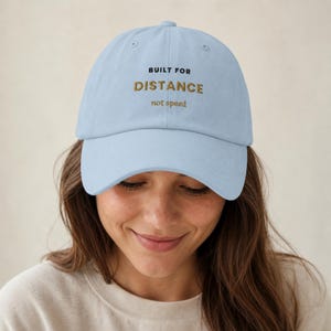 May include: A light blue baseball cap featuring embroidered text on the front. The text reads BUILT FOR in black, followed by DISTANCE in gold, and not speed in smaller gold lettering. The cap has a curved brim and a structured six-panel design.