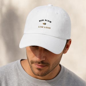 May include: A white baseball cap featuring the embroidered text RUN SLOW in black, a small gold megaphone icon, and the text LIVE LOUD in gold. The cap has a structured crown, a curved brim, and a button on top, providing a clean and simple look.