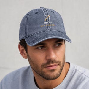 May include: A navy blue baseball cap with a distressed, washed-out texture. The front features an embroidered white circle with a diagonal gold line, accompanied by the white text MILES and the gold text NOT MINUTES below the graphic.