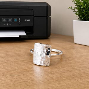 May include: A silver-toned ring featuring a large, rectangular centrepiece with a hammered, textured finish. The band is smooth and circular, connecting to the sides of the rectangular focal point, creating a reflective and metallic appearance.